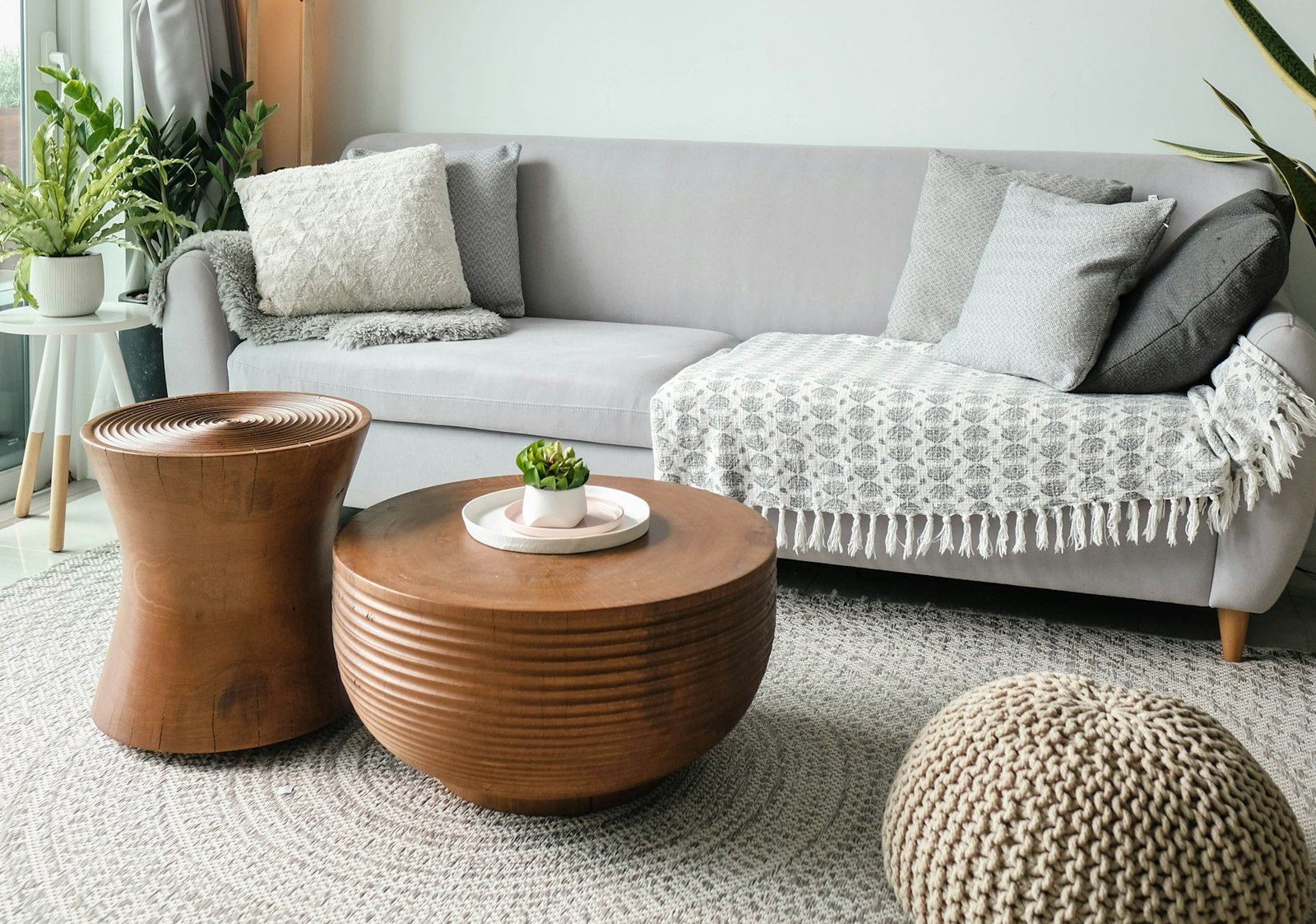 A bright consulting room with a soft grey sofa, wooden side tables, potted plants and daylight from a tall window